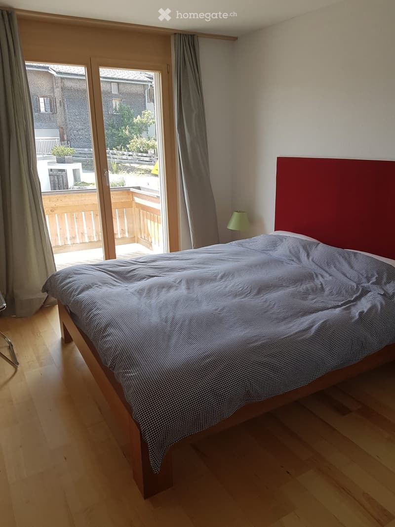 Cozy bedroom with wooden flooring, a neatly made bed with a checkered duvet, a red headboard, and a small green lamp on a bedside table. Large sliding glass doors lead to a balcony with outdoor seating.
