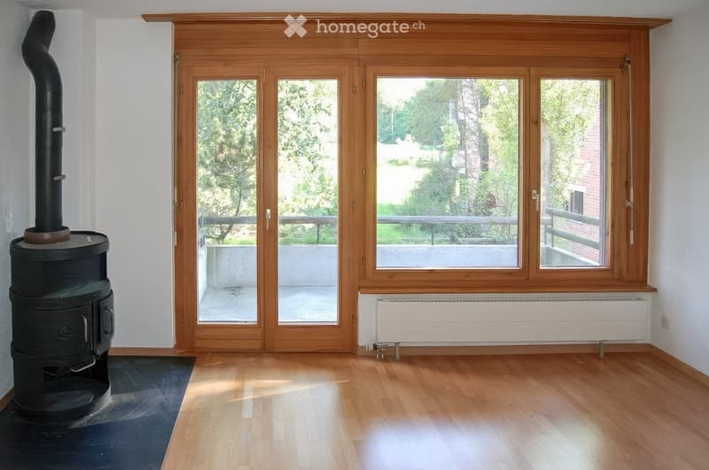 Empty room with wooden flooring, large wooden-framed windows, and a black wood-burning stove. The room has white walls and a radiator below the windows.
