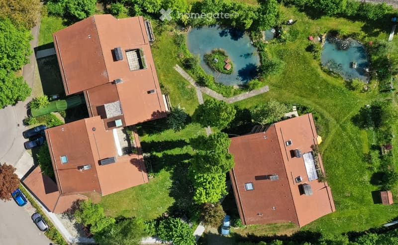 Aerial view of a residential house with a red-tiled roof, surrounded by a well-maintained garden, a pond, and several parked cars.