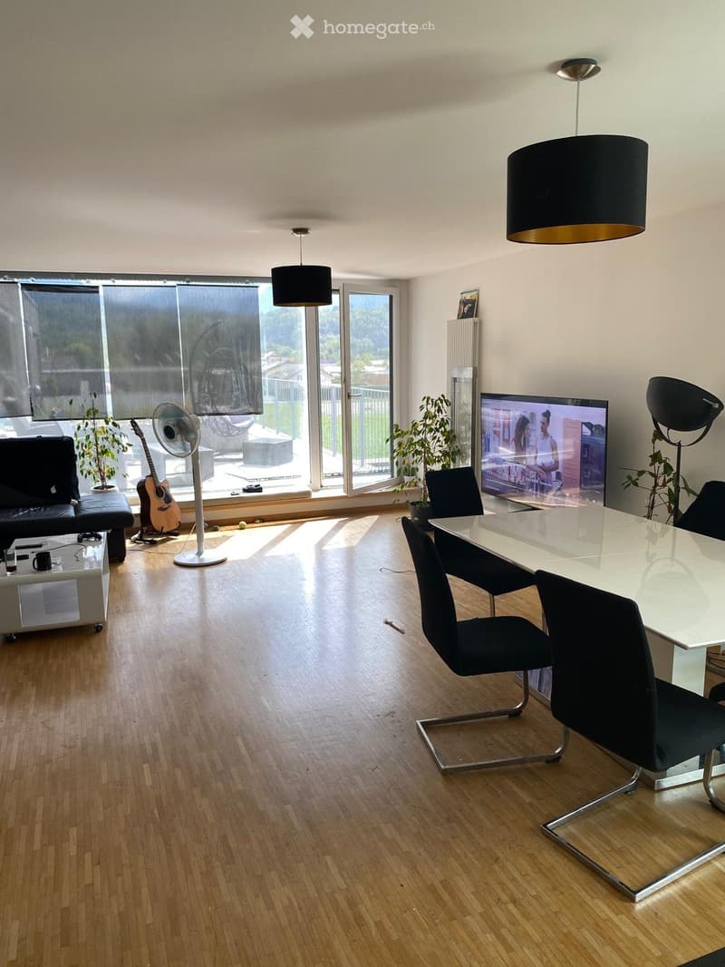 Modern living room with wooden flooring, large windows, a sofa, a coffee table, a TV, a guitar, a fan, and a dining area with a table and chairs.