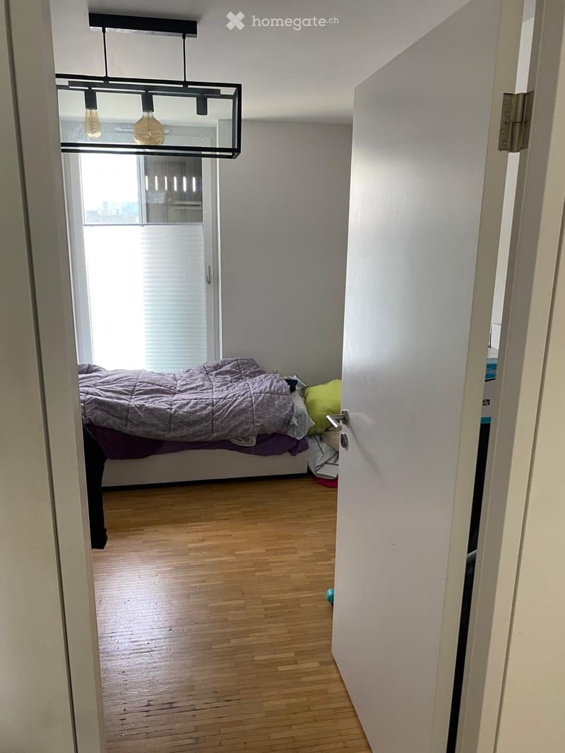 A small, well-lit bedroom with wooden flooring, a bed with a grey duvet and pillows, and a modern pendant light fixture above.