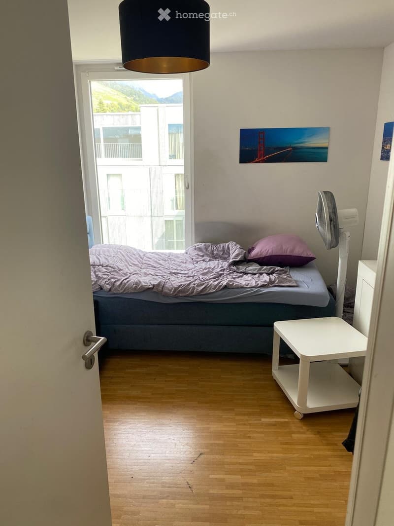 Cozy bedroom with wooden flooring, a double bed with grey bedding, a white nightstand, a fan, and a small white table near the door.