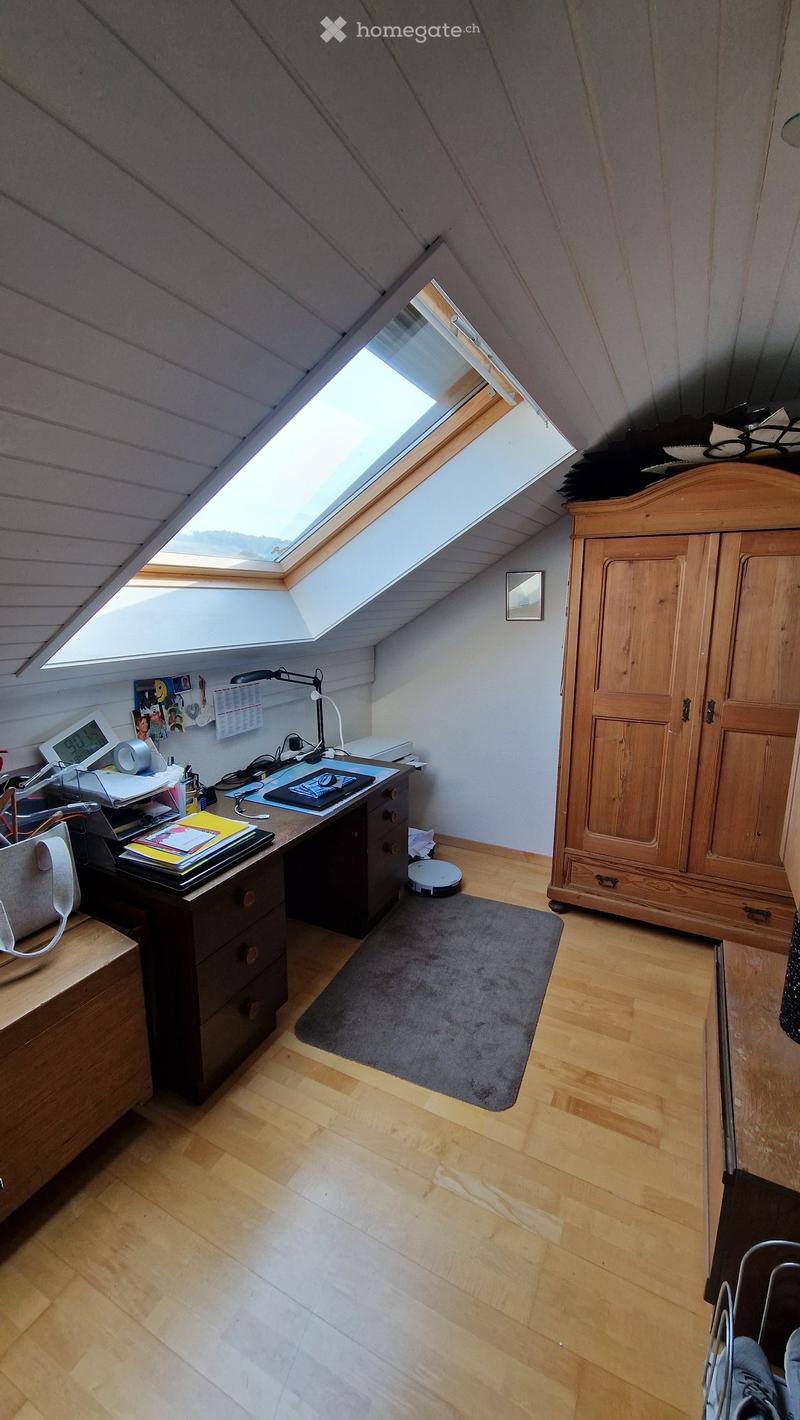 Bright attic room with wooden floors, slanted ceilings, and a skylight window. Features a wooden desk with a laptop, books, and a lamp, a large wardrobe, and a small gray rug.