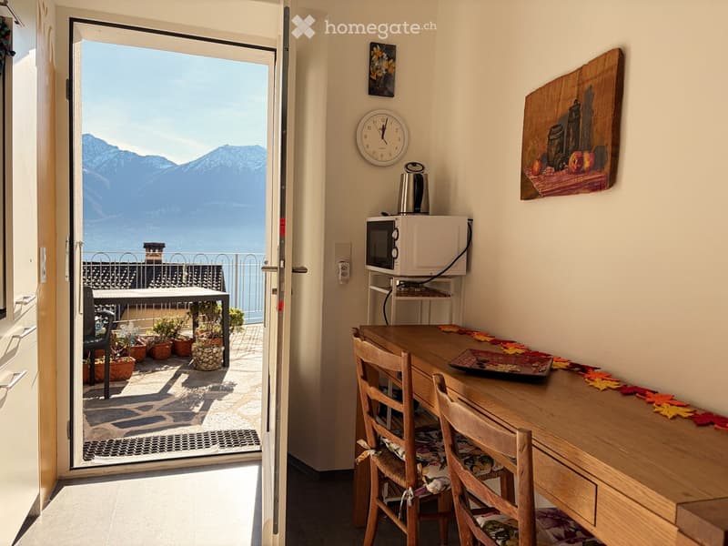 Bright room with a wooden table, two chairs, microwave, clock, and a view of mountains through an open sliding door leading to a balcony with potted plants.