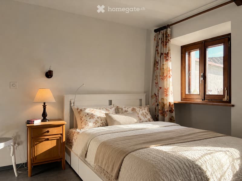Cozy bedroom with a white bed, patterned pillows, a nightstand with a lamp, and a window with floral curtains.