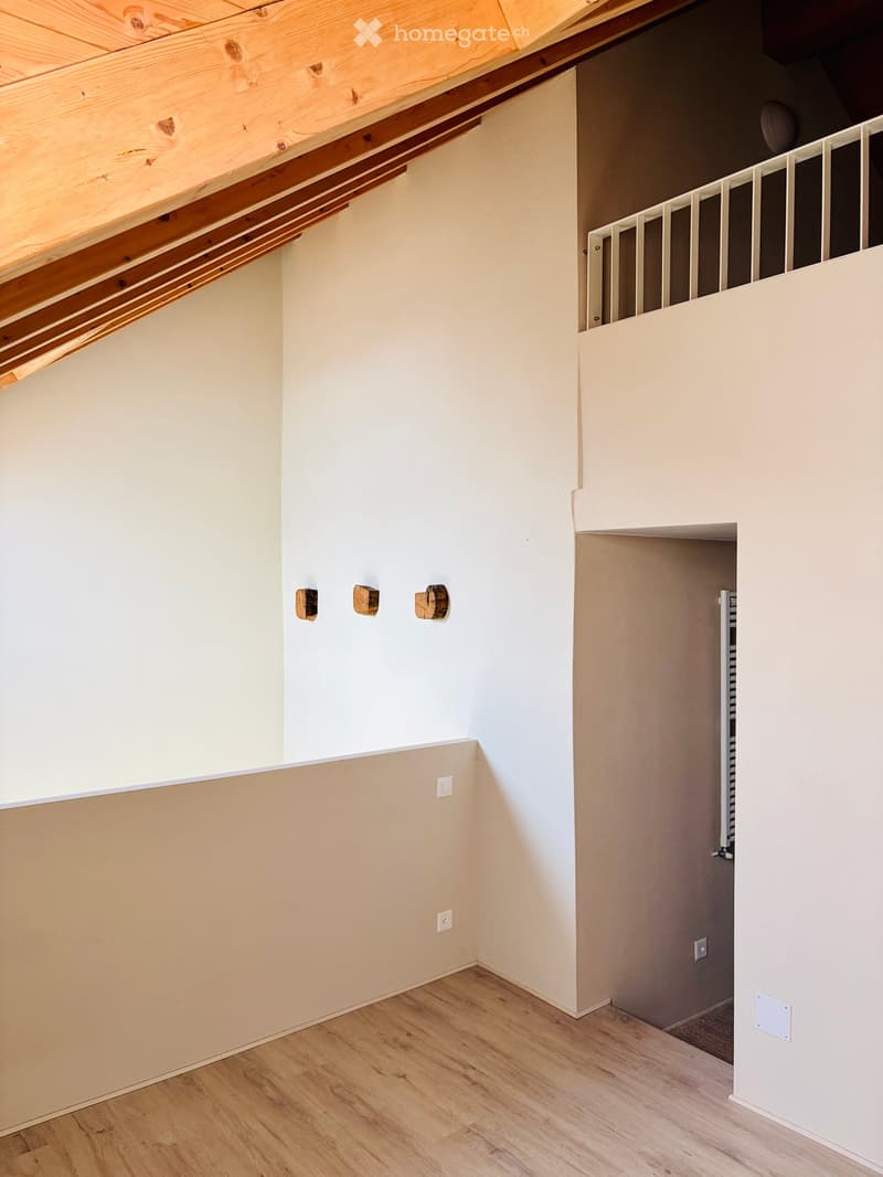 Empty room with light wooden flooring, white walls, and exposed wooden beams on the ceiling. A staircase with a white railing is visible in the background.