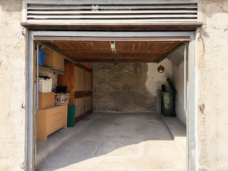A small, enclosed storage area with wooden cabinets, storage boxes, a basket, and a green trash bin. The space has a concrete floor and is accessed through a metal door.