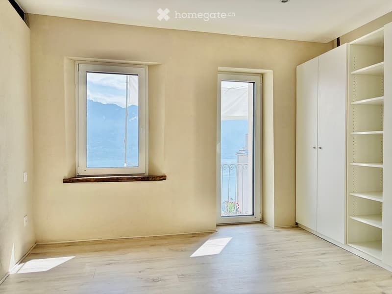 Empty room with light-colored walls, wooden flooring, a window with a scenic view, a door leading to a balcony, and built-in white shelving units.
