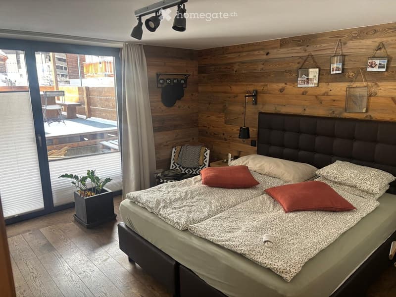 Modern bedroom with wooden walls, large bed with patterned bedding, nightstand, chair, and a small plant near the window. The room features a balcony with outdoor furniture visible through the large window.