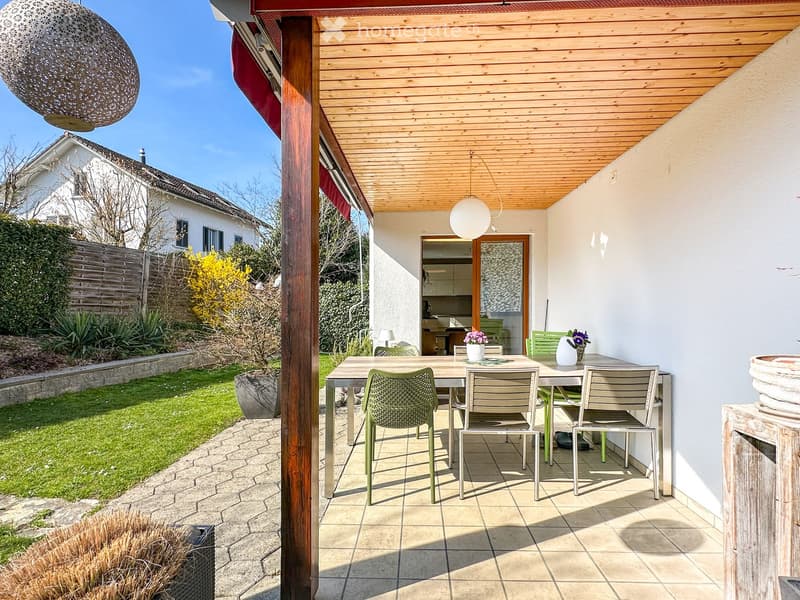 Outdoor patio with a wooden pergola, tiled floor, dining table with chairs, and a view of a well-maintained garden.