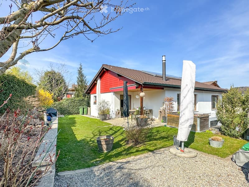 Well-maintained garden with a lawn, patio seating area, outdoor umbrella, and surrounding greenery.