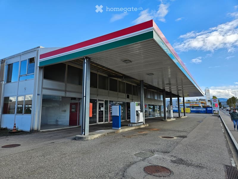 Modern gas station with a red and green roof, glass front, and fuel pumps under a covered area.