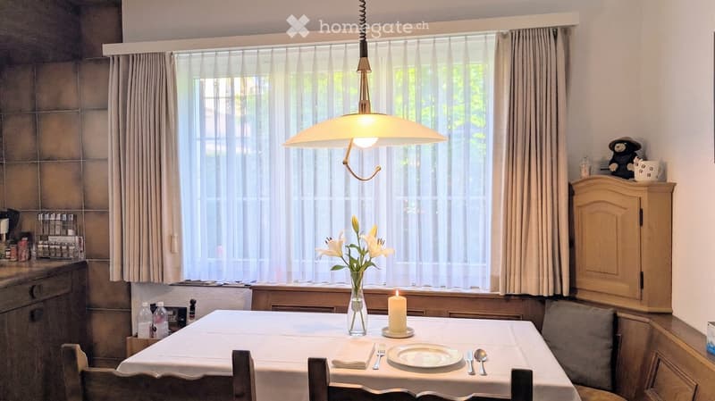 Dining room with a table set for two, a hanging lamp, a vase with flowers, and a small cabinet with a decorative item on top.
