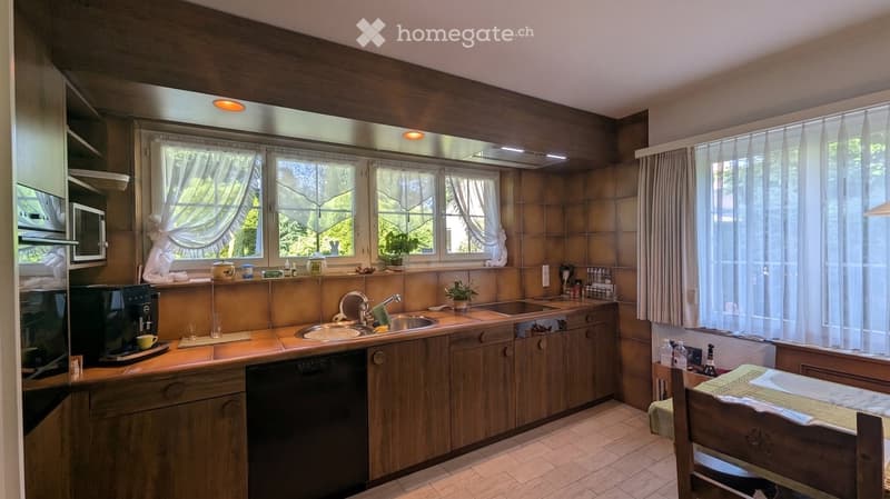A cozy kitchen with wooden cabinets, a central island, and a sink area. The space features a microwave, coffee maker, and a small dining area with a table and chair. Natural light streams in through large windows with curtains.