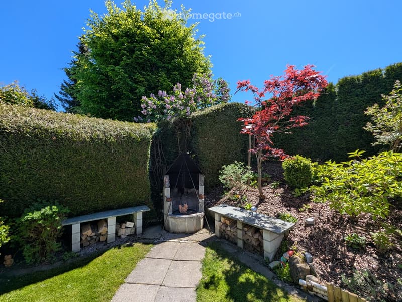 Well-maintained garden with a stone fireplace, neatly stacked firewood, benches, and various trees and shrubs.