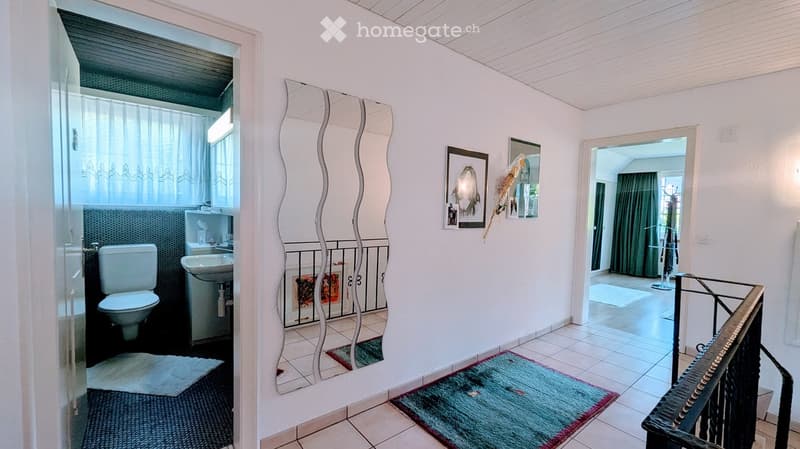 A well-lit hallway with white walls, tiled flooring, and a decorative mirror. It features a small rug, a staircase leading upwards, and an open doorway to a bathroom area with a toilet and sink.