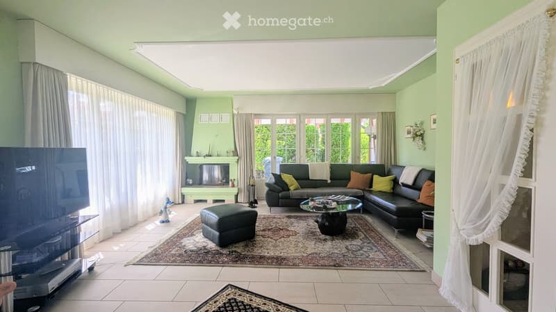 Bright living room with large windows, green walls, a black sectional sofa with colorful cushions, an ottoman, a glass coffee table, a fireplace, and a television on a stand.