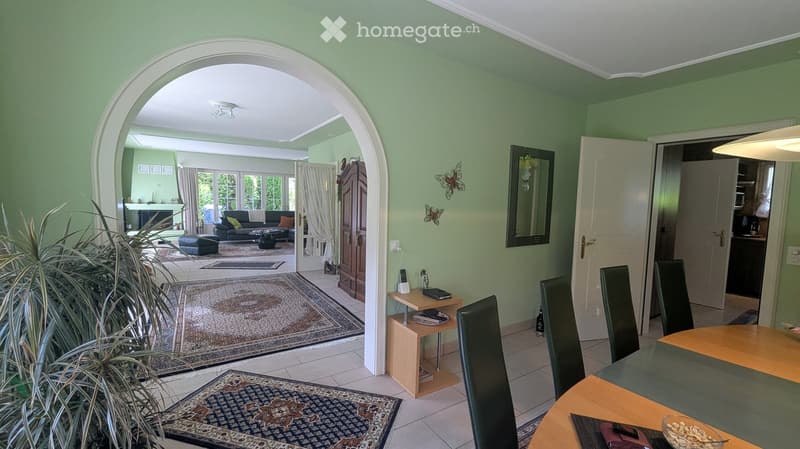 A well-lit living and dining area with light green walls, tiled flooring, and large windows. The living area features a sofa, coffee table, ottoman, and a small side table with a lamp. The dining area includes a wooden table with chairs and a decorative b