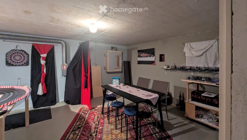The room features a table with chairs and stools, a shelf with various items, a dartboard on the wall, a toy race track, and a curtain separating part of the space.