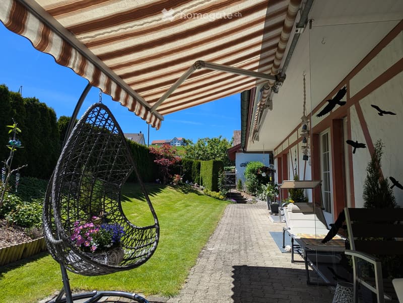 Garden with a covered patio, wicker hanging egg chair with flowers, lounge chairs, and a well-maintained lawn.