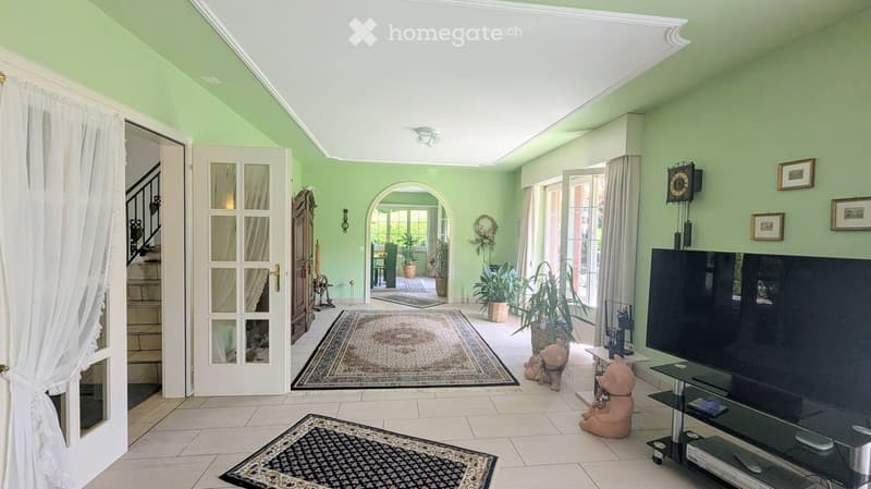 Bright living room with light green walls, white ceiling, tiled floor, large windows, television, decorative rugs, teddy bears, and framed pictures on the wall.