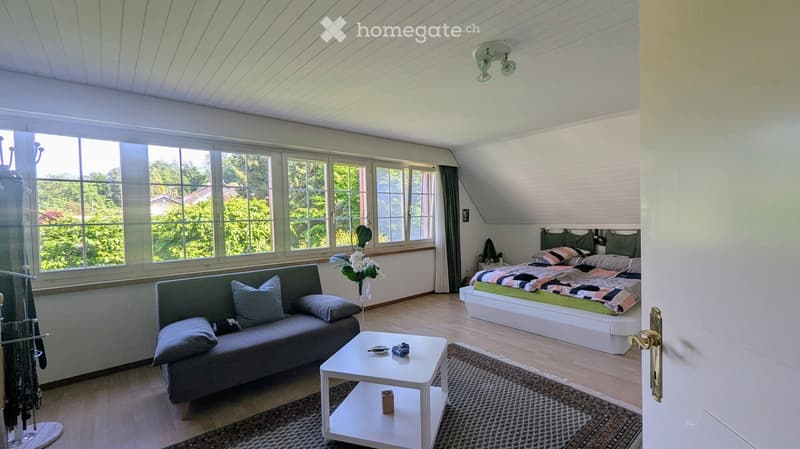 Bright, modern living space with a sofa, coffee table, bed, nightstand, and a flower vase. Large windows offer a view of greenery outside.