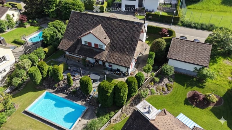 Aerial view of a two-story house with a well-maintained garden, a swimming pool, and surrounding greenery.