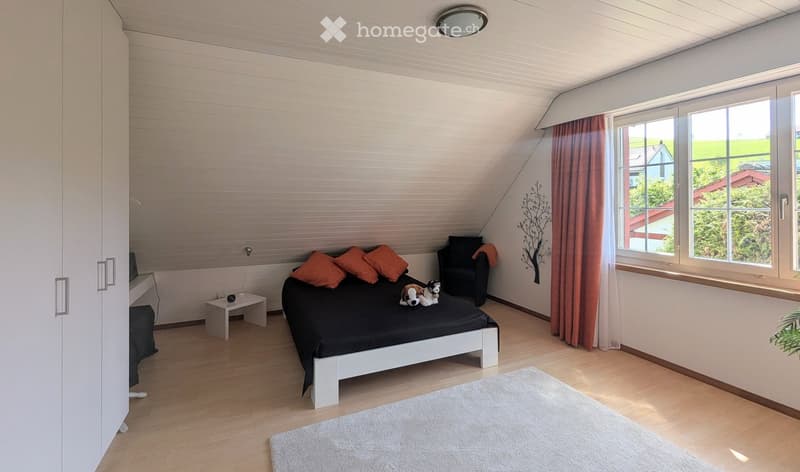 Bright bedroom with wooden flooring, large windows, a white bed with orange pillows, a nightstand, a small chair, and a decorative wall art piece.