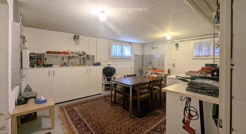 The room is a well-lit, multi-functional space with a dining area, kitchen cabinets, and a workbench area. It features a wooden dining table with chairs, a small stool, a refrigerator, and various tools and equipment neatly arranged on shelves and the wor