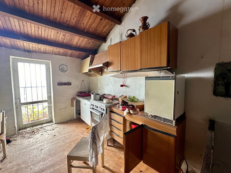 Small kitchen with wooden cabinets, a refrigerator, an oven, and a countertop with various items. The room has a slanted wooden ceiling and a window with bars.