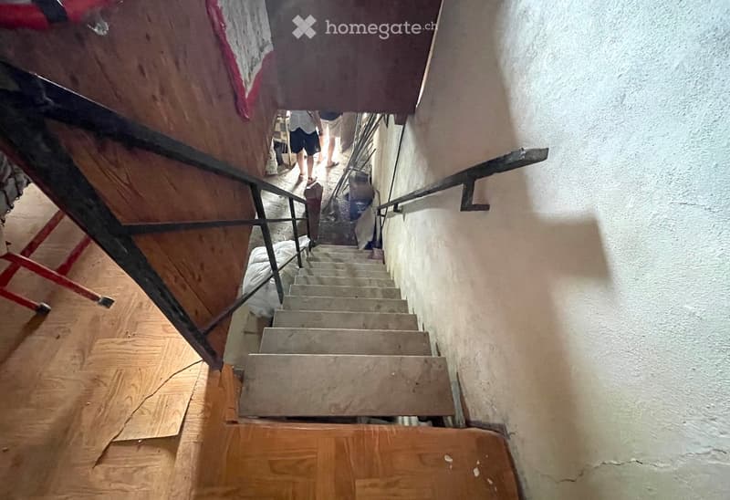 A narrow staircase with wooden steps and metal railings, leading upwards. The walls are plain and white, and the floor is covered with wooden planks. There are some people visible in the background, and a red ladder is partially visible on the left side.