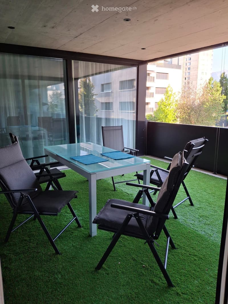Balcony with artificial grass, glass table, and several folding chairs.