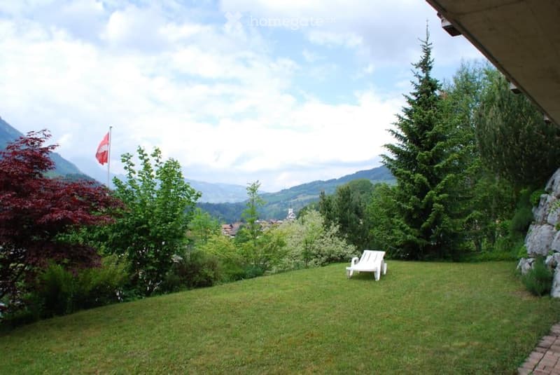 Lush green garden with a white lounge chair, surrounded by trees and a scenic mountain view.