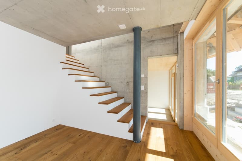 Modern hallway with wooden flooring, white concrete walls, and a staircase with wooden steps leading upwards. Features large windows and a central black column.