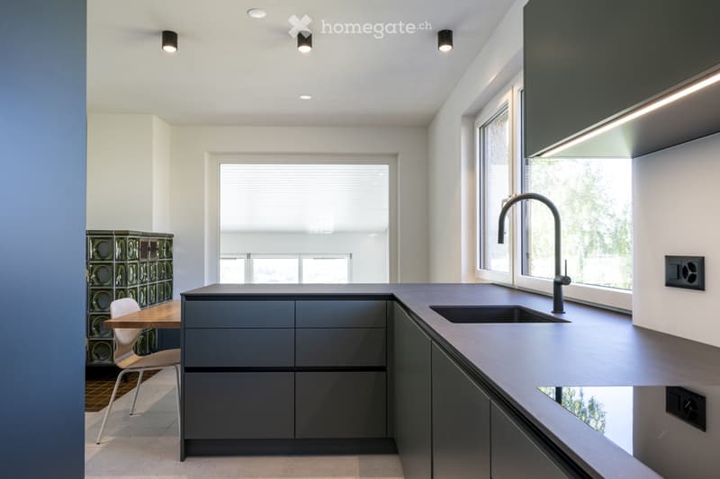 Modern kitchen with grey cabinets, a central island, a sink with a modern faucet, a small table with a chair, and a decorative glass block wall.
