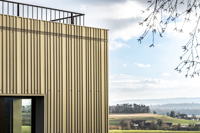 Modern building with vertical striped facade and balcony railing, overlooking a rural landscape with fields and scattered trees.