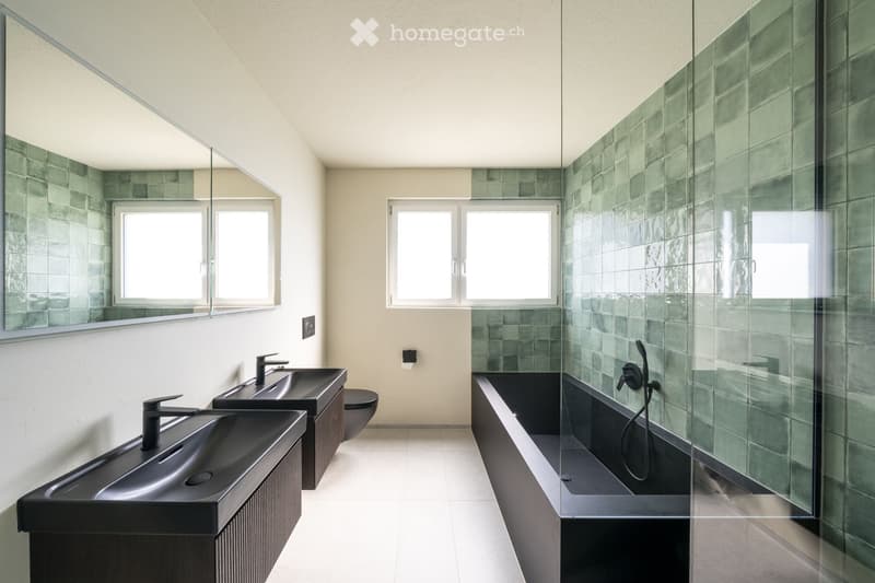 Modern bathroom with two black sinks, a large mirror, a bathtub with glass enclosure, and a toilet. Green tiled walls and white flooring.