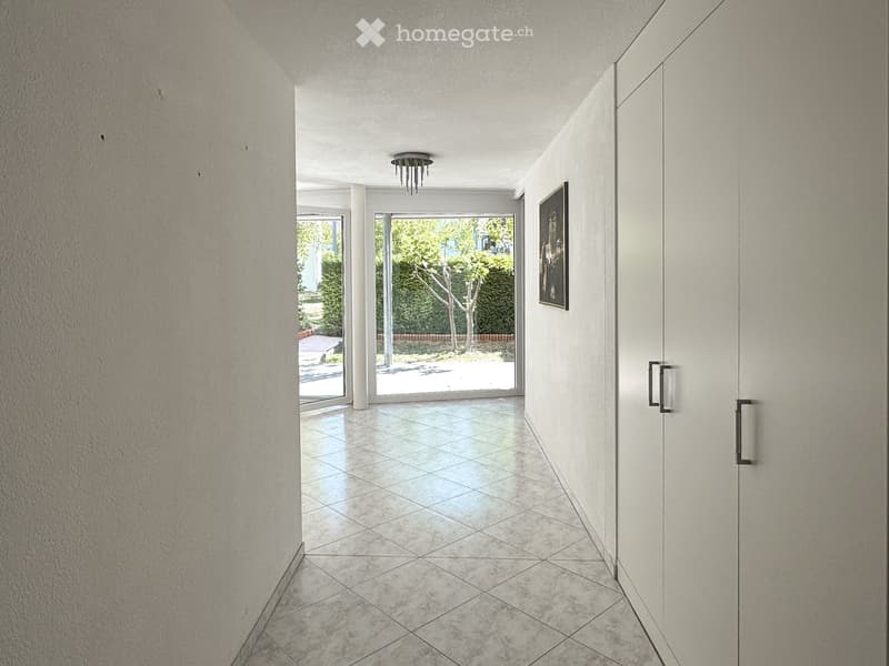 Modern hallway with tiled floor, white walls, recessed lighting, and a framed picture on the wall. The hallway leads to a bright outdoor area with greenery visible through large glass doors.
