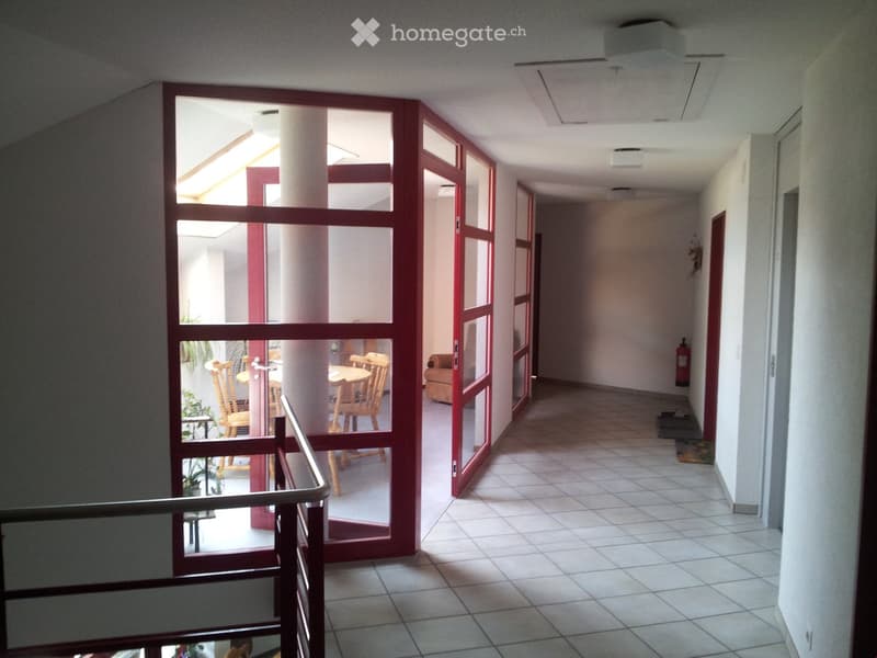 A well-lit hallway with tiled flooring, white walls, and red-framed glass doors leading to a room with wooden chairs and a table. A fire extinguisher is mounted on the wall.