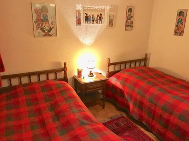 Cozy bedroom with two single beds, red and green checkered bedding, a wooden nightstand with a lamp, and several picture frames on the walls.