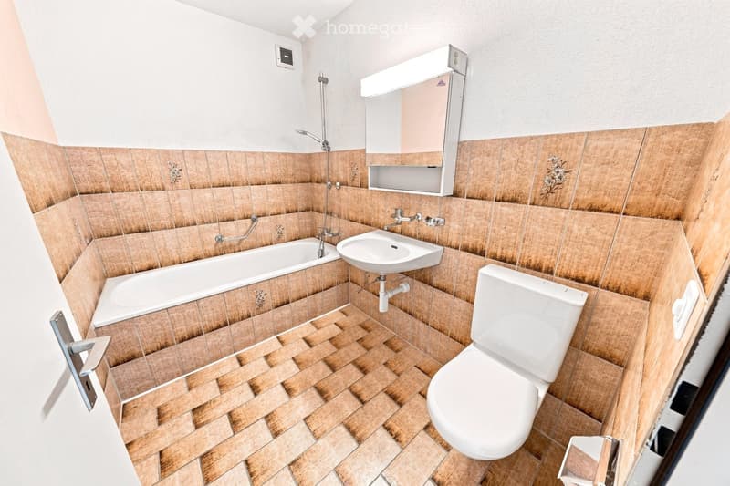 Bathroom with a bathtub, sink, toilet, and mirror. The walls are tiled in a warm, wood-like pattern, and the floor has a herringbone tile design.