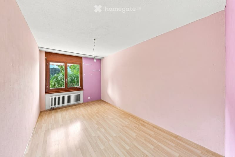 Empty room with light pink walls, wooden flooring, and a window with wooden frame. There is a radiator below the window and a door leading to another room.