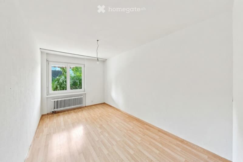 Empty room with light wooden flooring, white walls, and a single window with a radiator below it.