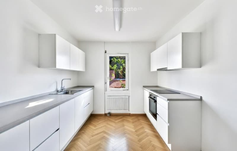 Modern, minimalist kitchen with white cabinets, grey countertops, and a wooden herringbone floor. Features a sink, faucet, and oven.