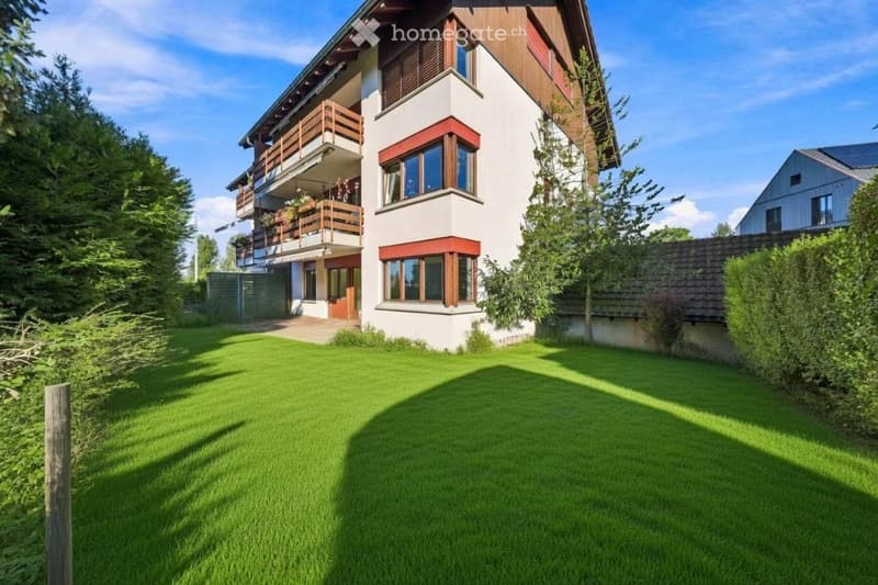 A two-story house with white walls, wooden accents, and large windows. Features include a balcony on the upper floor, a well-maintained lawn, and surrounding trees.