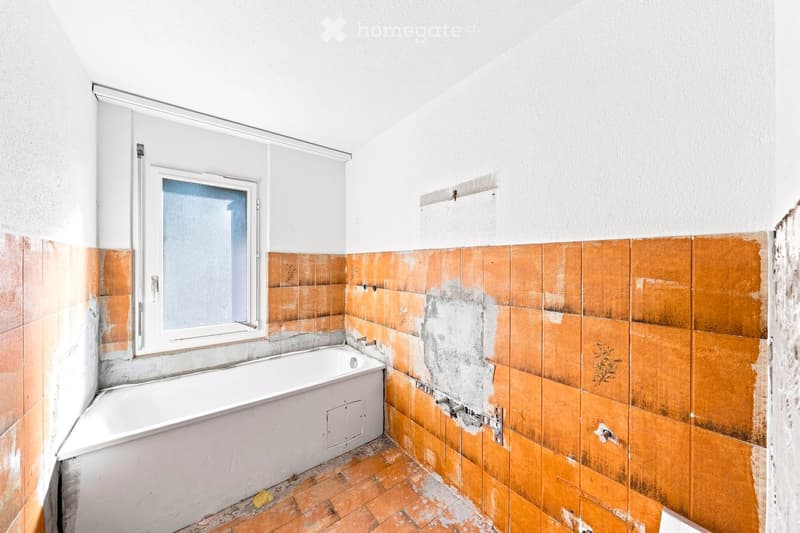 Bathroom with a white bathtub, orange tiles, and a window with frosted glass. The tiles show signs of wear and discoloration.