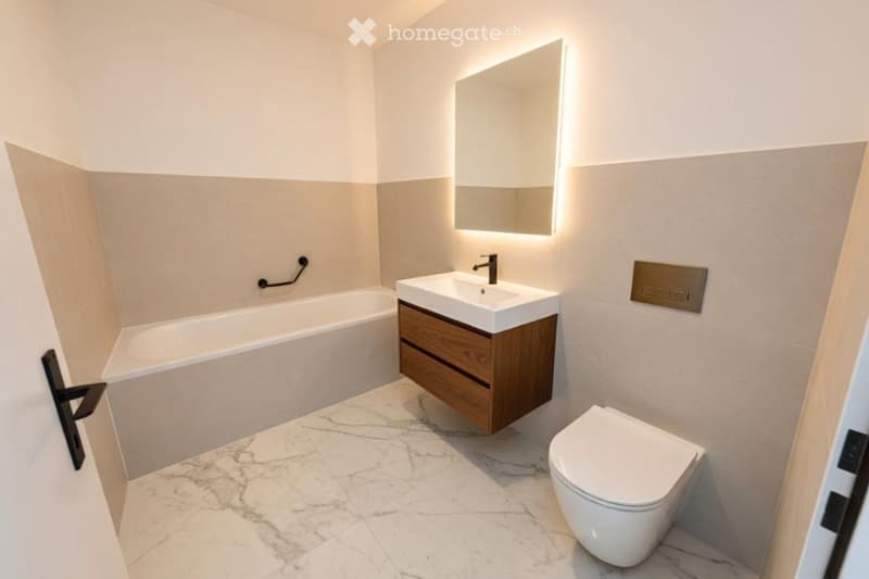 Modern bathroom with a white bathtub, a wooden vanity with a sink, a toilet, and a large illuminated mirror above the sink. The floor is covered with marble tiles.