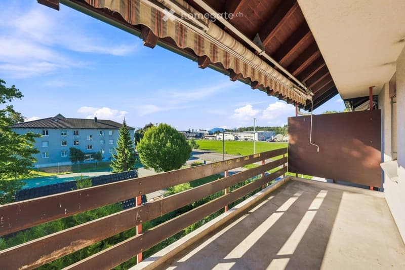 Balcony with wooden railing, striped awning, and scenic view of a green field, trees, and distant buildings.