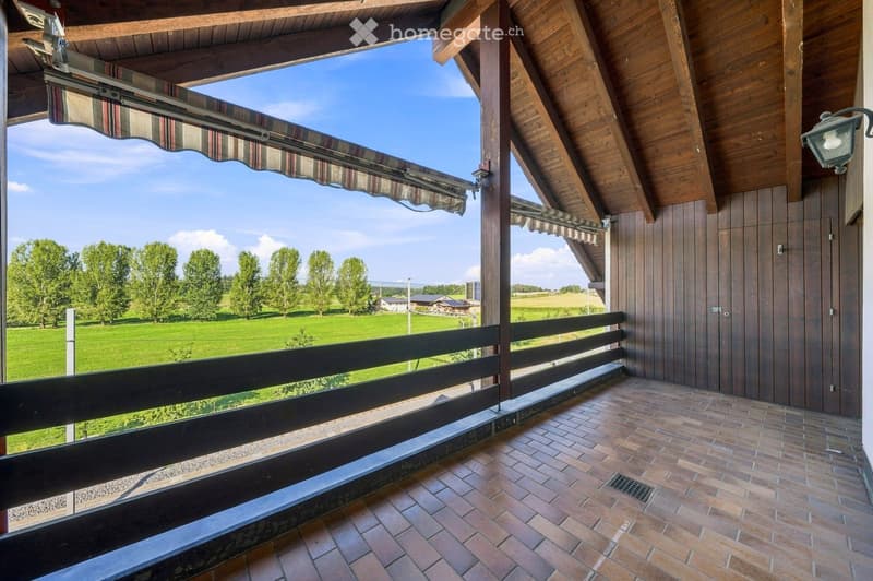 Balcony with wooden beams, brick tiles, and a view of a green field and trees.