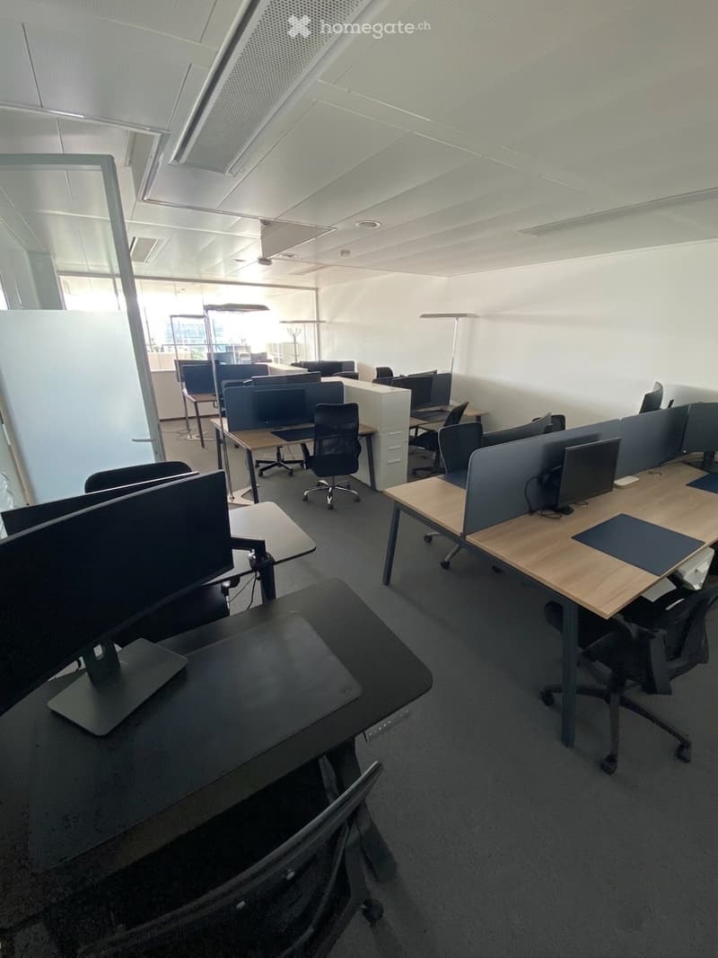 Modern office space with multiple desks, chairs, computer monitors, and screen partitions. The room features a clean, minimalist design with white walls and a gray carpeted floor.
